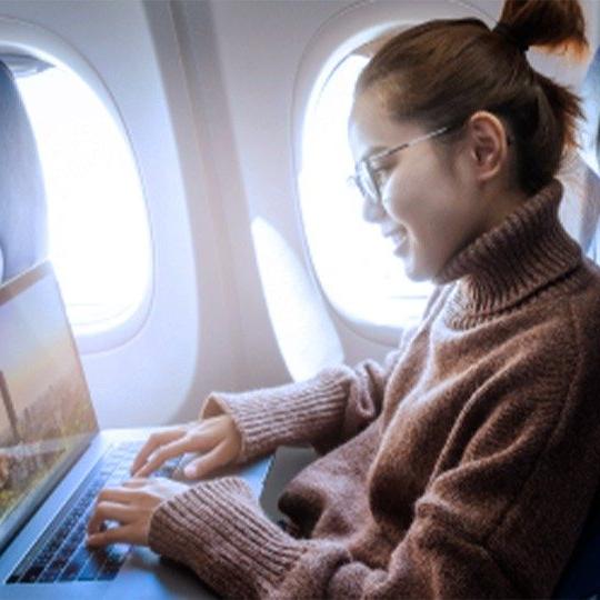 Woman wearing glasses and a brown sweater is working during a flight on her laptop over onbaord WiFi