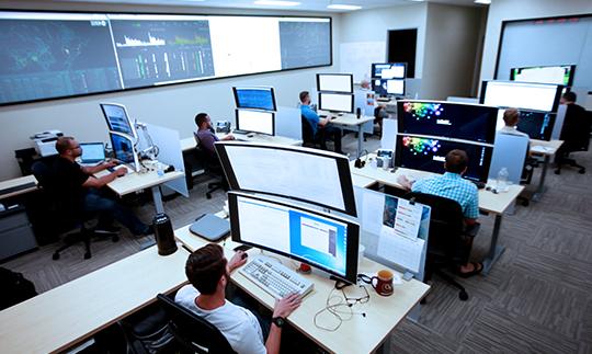 Viasat员工在2张大桌子上工作, 网络安全运营中心的堆叠显示器